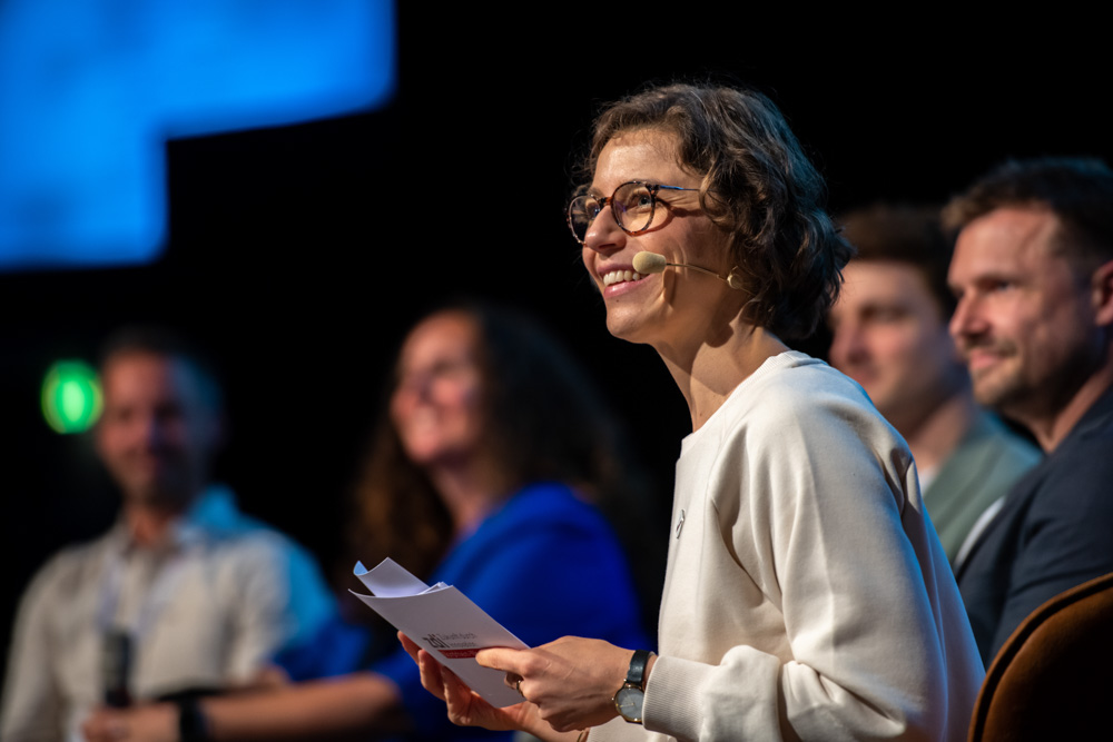 Eine Frau mit Brille und Headset-Mikrofon hält Papiere in der Hand und spricht auf der Bühne. Vier sitzende Personen sind unscharf hinter ihr zu erkennen.