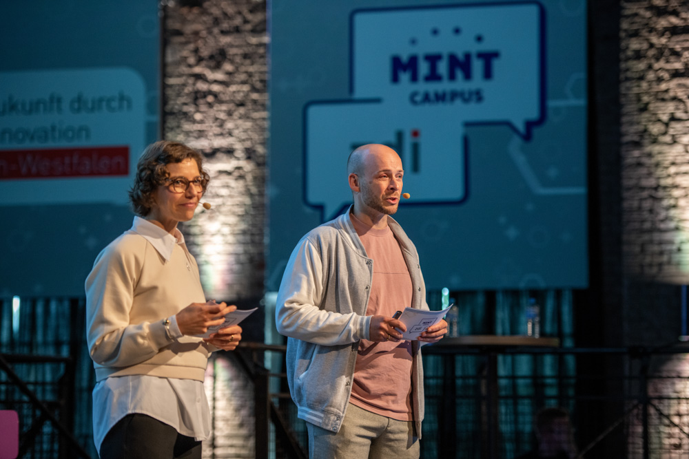 Zwei Personen stehen auf der Bühne und halten Moderationskarten in der Hand. Im Hintergrund sind die Logos von "MINT-Campus" und "zdi.NRW" zu sehen.