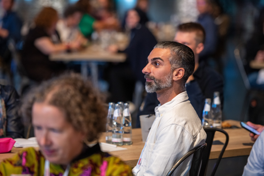 Ein Mann in einem weißen Hemd sitzt an einem Tisch und blickt aufmerksam nach vorne. Andere Menschen sitzen in der Nähe in einem belebten Innenraum und haben Wasserflaschen auf dem Tisch stehen.
