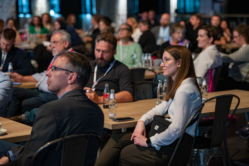 Menschen sitzen an Tischen in einer Halle und hören einer Person auf der Bühne zu.