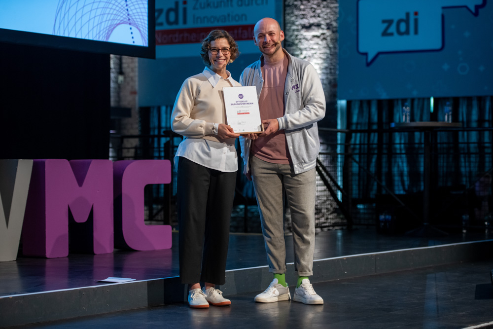 Zwei Personen stehen auf einer Bühne und halten eine Urkunde in der Hand.