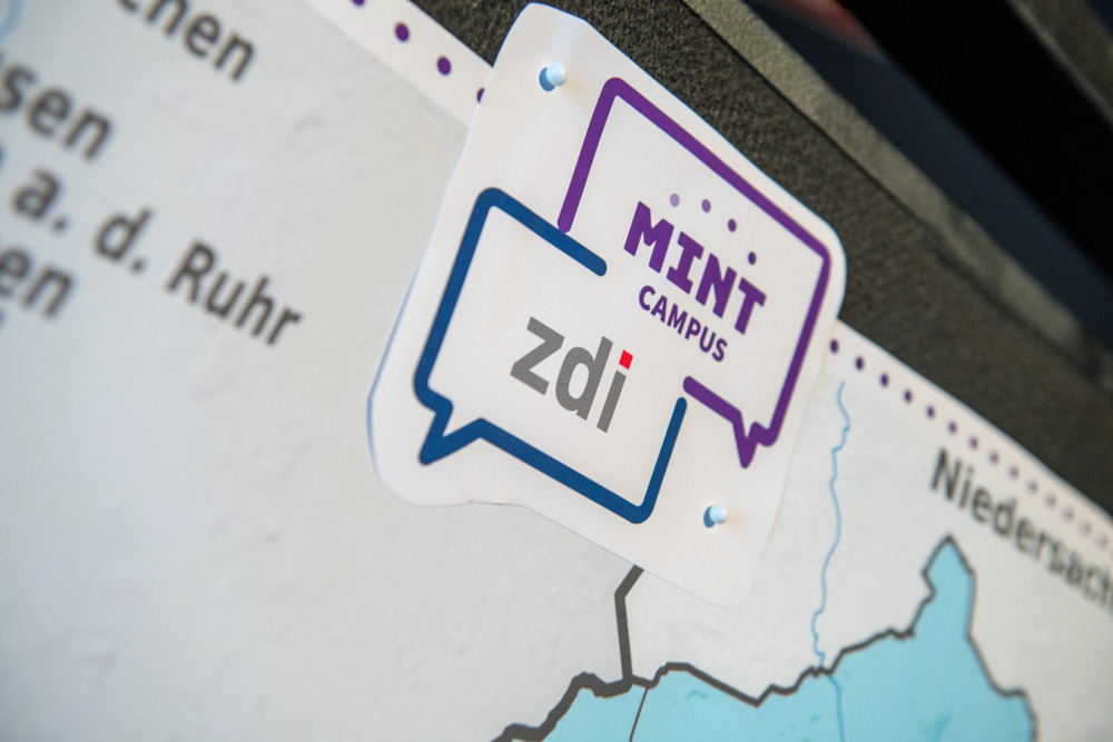 Das Bild zeigt eine Nahaufnahme einer Landkarte. Ein angeheftetes Schild zeigt in Sprechblasen die Logos "MINT-Campus" und "zdi.NRW".