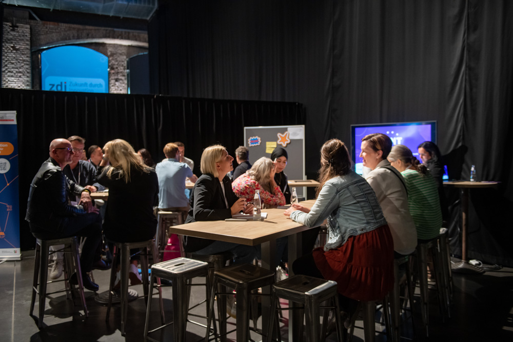 Mehrere Personen sitzen und unterhalten sich an Stehtischen in einem Veranstaltungsraum.