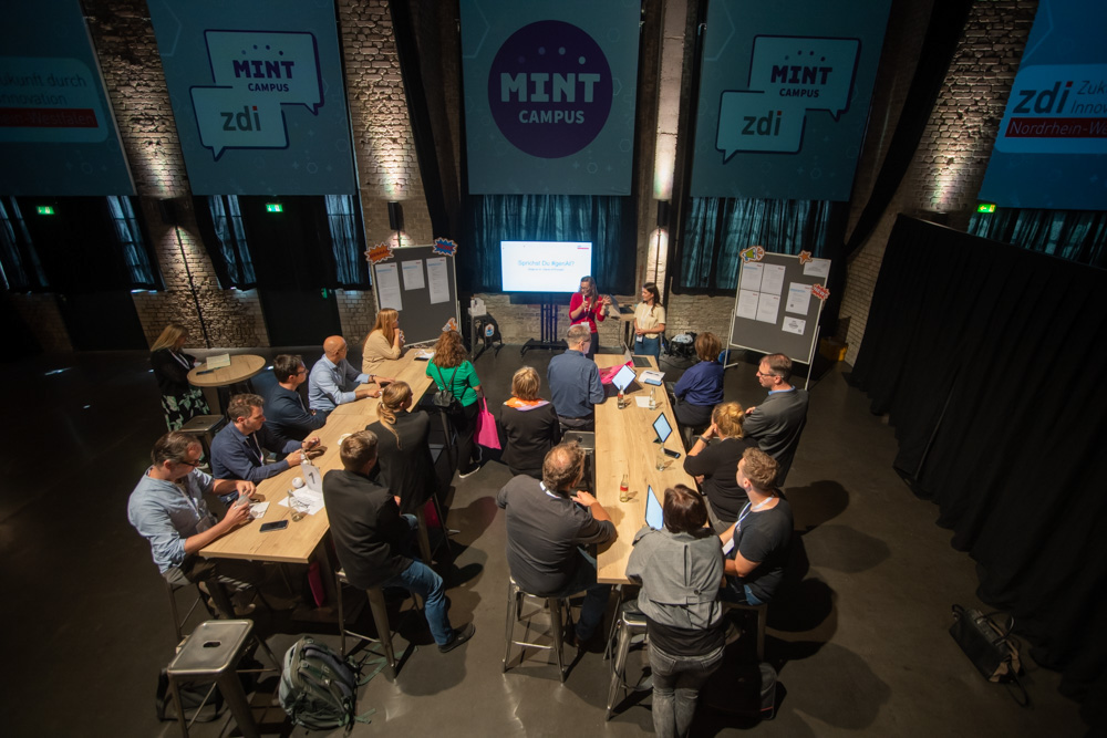 Menschen sitzen um zwei lange Tische in einem Workshop. Sie sehen eine Präsentation auf einem Bildschirm.
