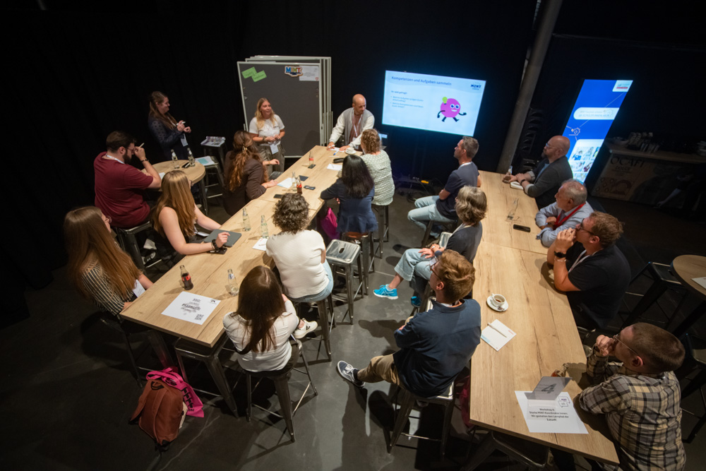 Menschen sitzen in zwei Gruppen an Tischen. Vor ihnen stehen ein Bildschirm, mehrere Pinnwände und die drei Dozent:innen.