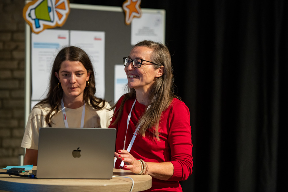 Zwei Frauen stehen an einem runden Tisch. Sie schauen auf einen Laptop und lächeln, während sie eine Präsentation halten.