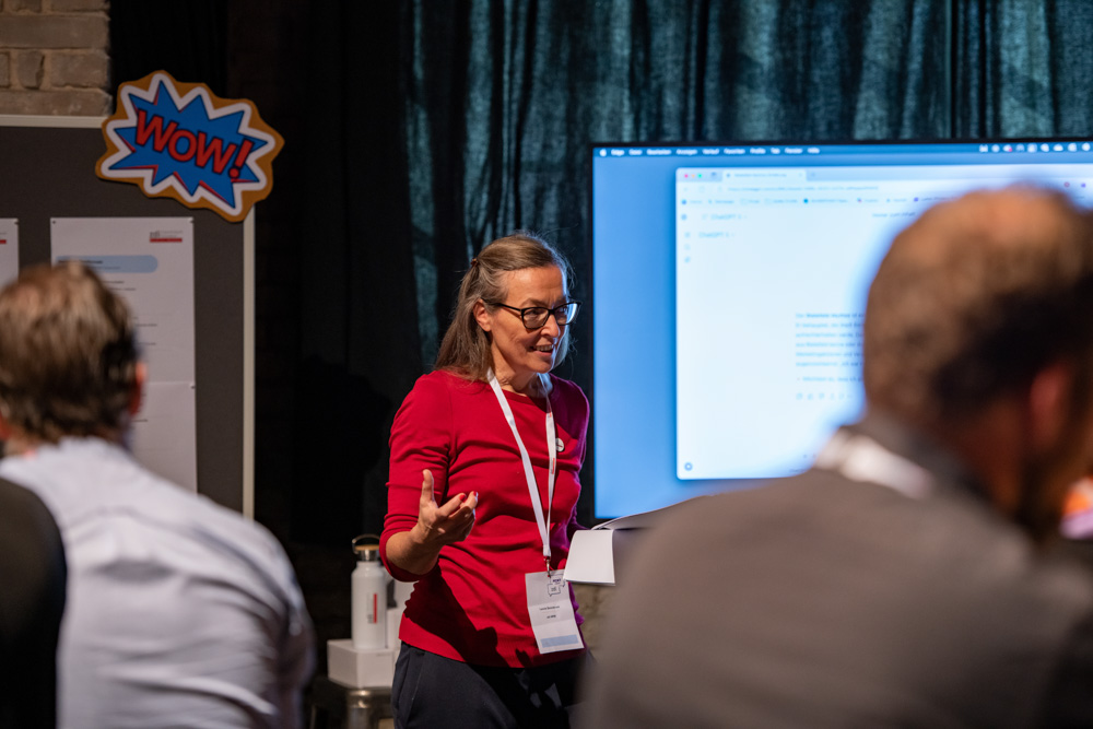 Eine Frau in einem roten Oberteil präsentiert sich einem Publikum vor einer Leinwand bei einer Indoor-Veranstaltung mit einem "WOW!"-Schild an der Wand.