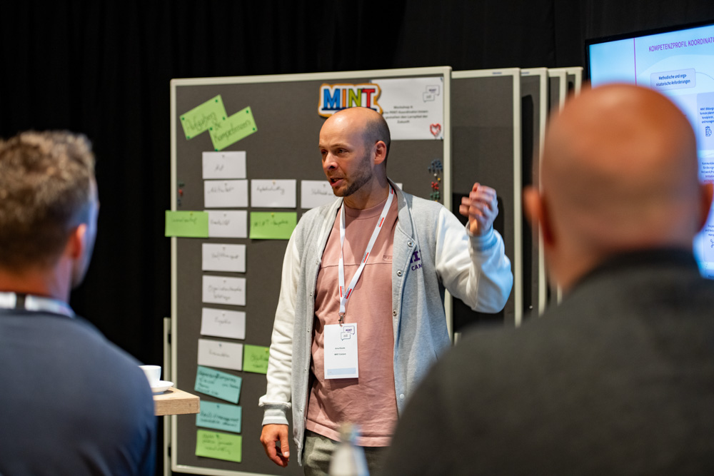 Ein Mann steht neben einem Bildschirm und präsentiert etwas. Mehrere Personen sitzen davor an Tischen und verfolgen die Präsentation.