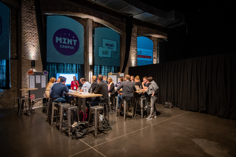 Das Bild zeigt eine Gruppe Menschen, die an zwei Tischen verteilet zusammen sitzen. Eine Frau steht vor einem Bildschirm und erzählt etwas. Im Hintergrund sieht man die Logos von zdi.NRW und MINT-Campus.