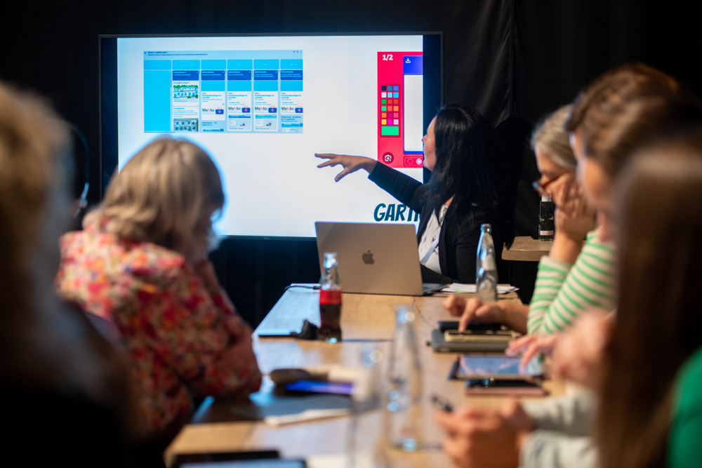 Eine Frau steht neben einem Bildschirm und präsentiert etwas. Mehrere Personen sitzen davor an Tischen und verfolgen die Präsentation.