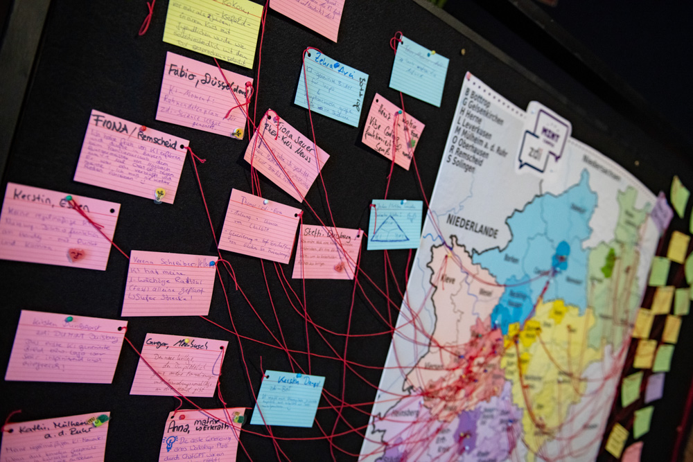 Das Bild zeigt eine Karte von Westdeutschland mit farbigen Notizen und roten Schnüren, die Orte miteinander verbinden. In der Nähe sind handgeschriebene Karten angeheftet.
