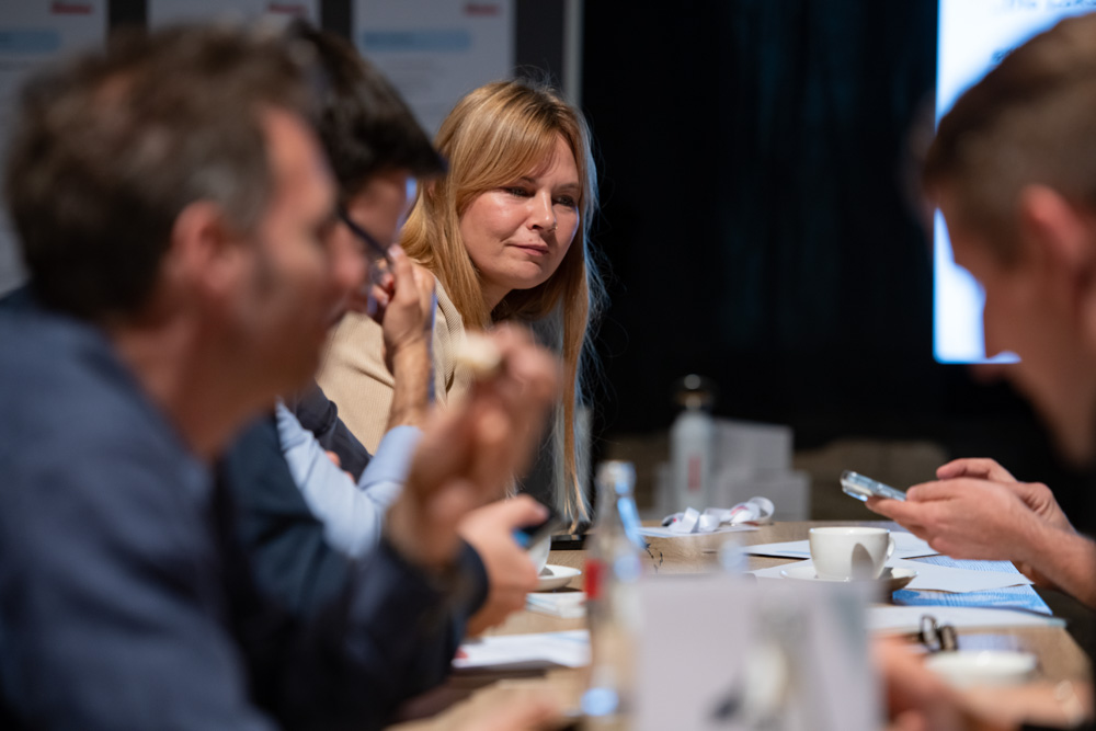 Eine Gruppe von Menschen sitzt an einem Tisch und diskutiert. Eine Frau steht im Mittelpunkt und schaut aufmerksam zu einer anderen Person.