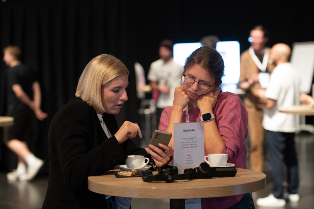 Zwei Frauen sitzen an einem hohen Tisch und schauen gemeinsam auf ein Smartphone. Auf dem Tisch stehen Kaffeetassen und eine Speisekarte, andere sind dahinter verschwommen.