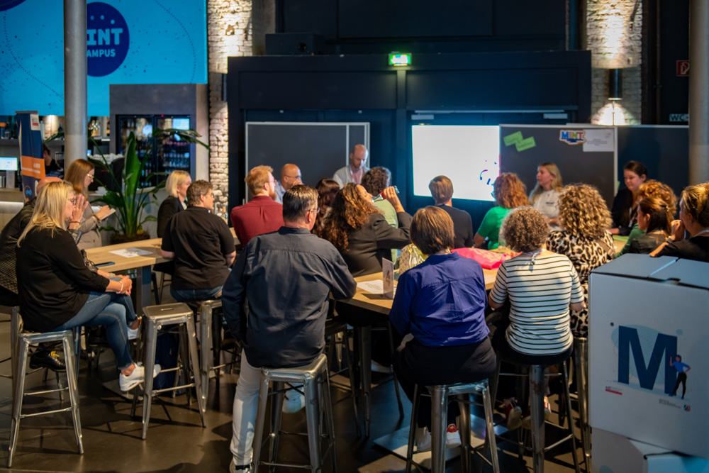 Die Menschen sitzen auf Hockern um Tische herum und hören einem Redner zu, der vor einem Bildschirm in einem Veranstaltungsraum steht.