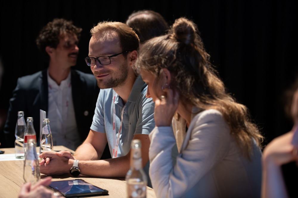 Zwei Personen sitzen an einem Tisch mit Getränken in Flaschen und unterhalten sich bei einer Veranstaltung in einem Gebäude, während andere Personen im Hintergrund zu sehen sind.