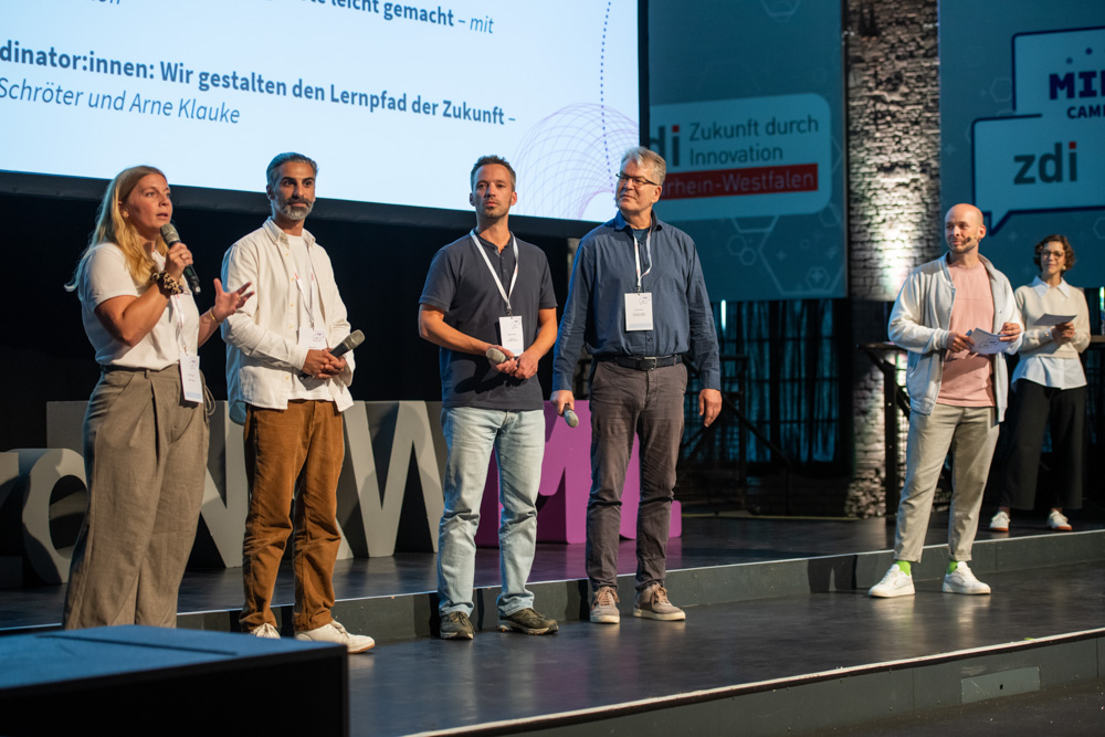 Menschen stehen auf der Bühne. Eine Frau spricht in ein Mikrofon, die anderen schauen sie an.