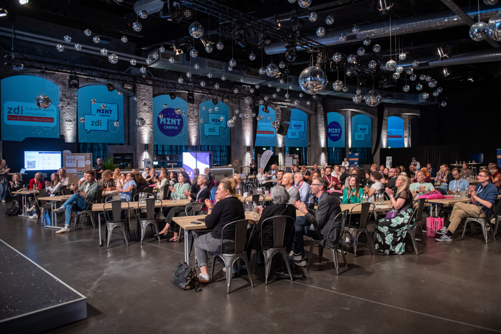 In einer geräumigen, mit Lichtern und Bannern geschmückten Veranstaltungshalle im Industriestil sitzen die Besucher an Tischen und vor einer Bühne.