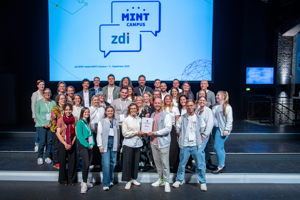 Das Foto zeigt eine Gruppe Menschen, die in die Kamera lächeln. Im Hintergrund sind die Logos von zdi.NRW und dem MINT-Campus zu sehen.