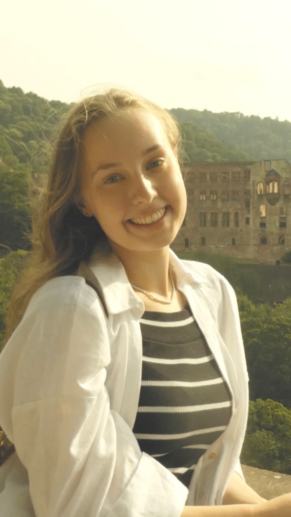 Das Foto zeigt Raja Flüchter, eine Frau mit langen Haaren, die in die Kamera lächelt. Im Hintergrund sind Bäume und ein altes Gebäude zu sehen.