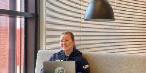 Eine Person sitzt auf einem beigen Sofa mit einem Laptop auf dem Schoß und lächelt. In der Nähe stehen ein kleiner weißer Tisch, eine schwarze Lampe und ein großes Fenster.