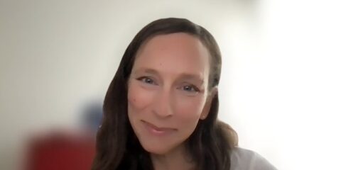 Die Frau hat langes braunes Haar und lächelt in die Kamera. Sie befindet sich in einem gut beleuchteten Raum mit einem unscharfen Hintergrund.