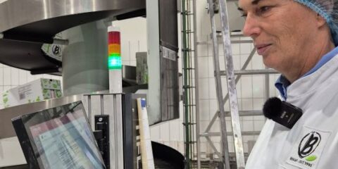 Eine Person im Laborkittel und mit Haarnetz benutzt einen Touchscreen-Computer zur Überwachung einer Produktionslinie in einer Industrieanlage.