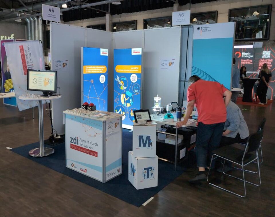 Ein Messestand zeigt Infotafeln, Broschüren und technische Geräte. Zwei Personen sitzen, eine benutzt einen Laptop. Werbeartikel liegen auf Tischen.