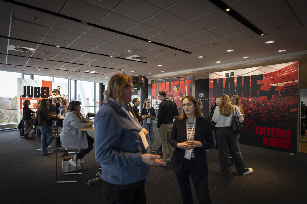 In einem modernen Veranstaltungsraum mit großen Wandgrafiken zum Thema Sport und Stehtischen unterhalten sich die Menschen. Andere stehen in kleinen Gruppen.