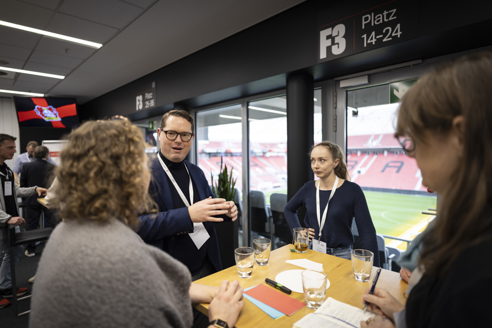 In einer Stadionsuite mit leeren Sitzen und einem Fußballfeld vor der Tür stehen Menschen um einen Tisch mit Getränken und Notizbüchern herum und unterhalten sich.