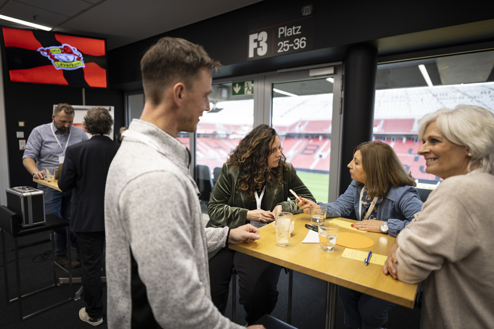 Die Leute unterhalten sich an einem hohen Tisch in einer Suite mit Getränken und Notizblöcken, mit Blick auf ein Fußballstadion.