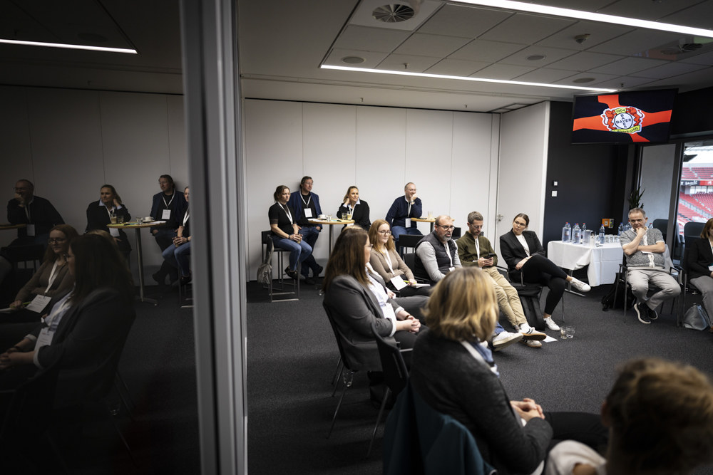Die Teilnehmer sitzen in einem Konferenzraum und schauen nach vorne. Hinter ihnen erscheint ein Logo auf einem Präsentationsbildschirm. Auf den Tischen stehen Getränke.