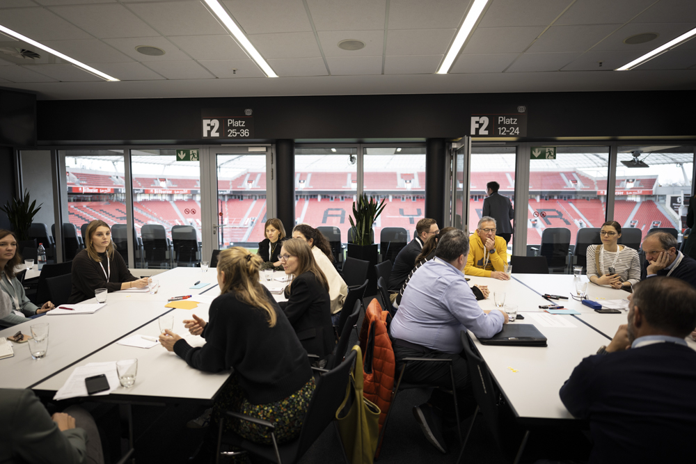 In einem Konferenzraum sitzen Menschen an Tischen und diskutieren. Große Fenster hinter ihnen zeigen ein Sportstadion im Freien.