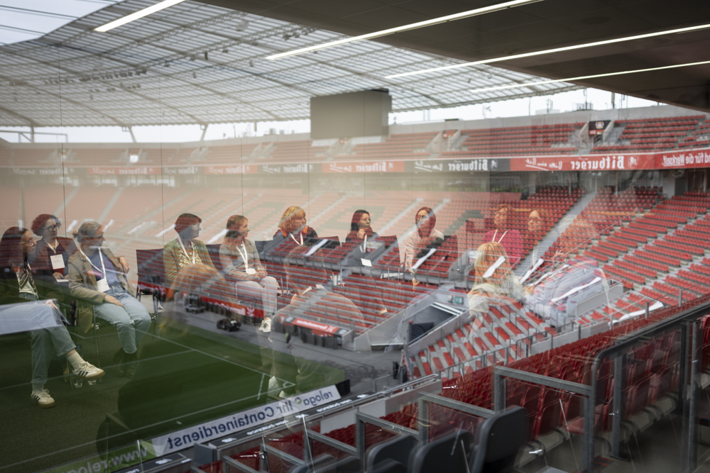 Menschen sitzen in einem Konferenzraum. Sie spiegeln sich in einem Fenster, das den Blick auf leere rote Sitzreihen im Stadion freigibt.