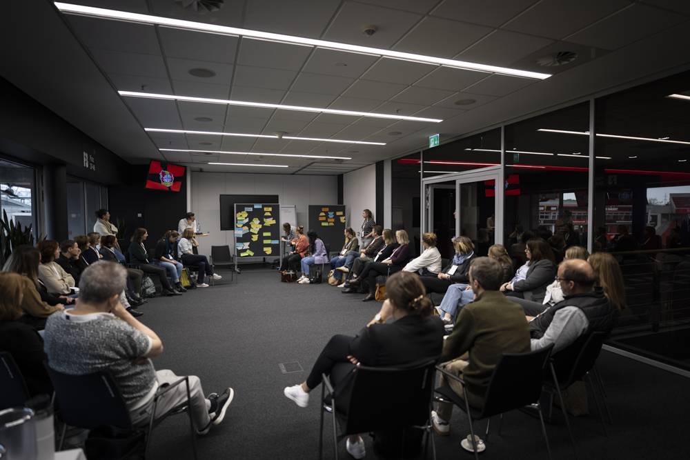 Die Leute sitzen im Halbkreis in einem Konferenzraum und hören einem Redner zu. Zwei Tafeln an der Stirnseite sind mit Haftnotizen beklebt.