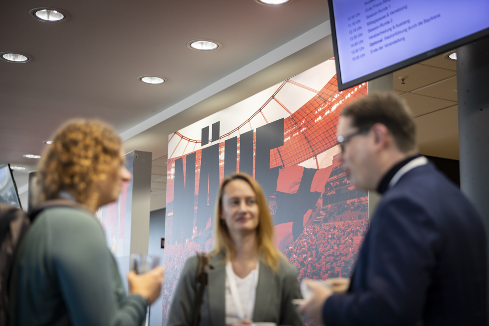 Drei Personen unterhalten sich in einem modernen Innenraum. Im Hintergrund sind eine große Wandmalerei und ein digitaler Bildschirm zu sehen.