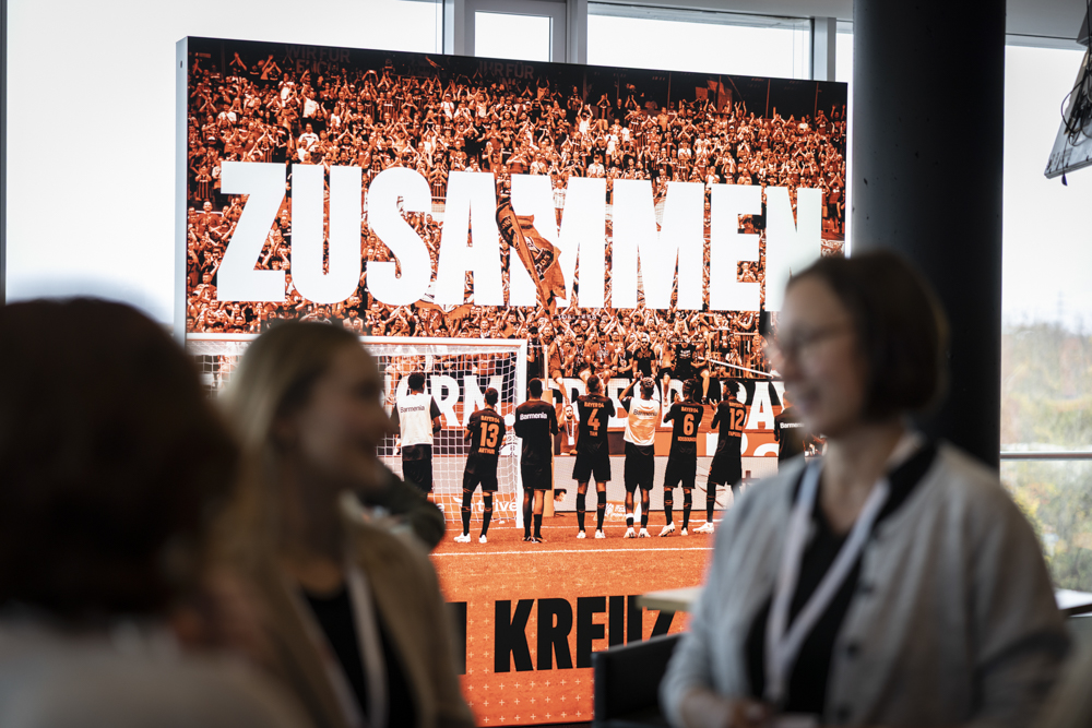 Ein großes Plakat mit der Aufschrift "ZUSAMMEN" ist hinter einer Fußballmannschaft zu sehen, die vor einer Menschenmenge feiert. Mehrere Personen stehen unscharf davor.