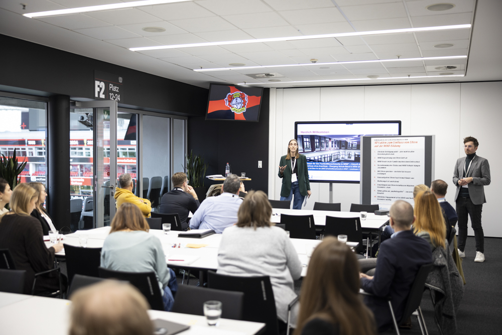 Eine Frau hält eine Präsentation vor einer sitzenden Gruppe in einem modernen Konferenzraum. Ein Mann steht in der Nähe, mit einem Bildschirm und einer Tafel an der Stirnseite.