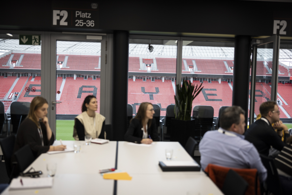 Menschen sitzen an einem Konferenztisch in einem Raum mit Blick auf ein leeres Stadion, dessen rote Sitze das Wort "BAYER" bilden.