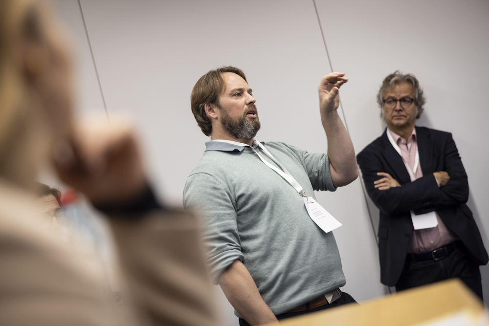 Ein Mann in einem grauen Pullover spricht und gestikuliert mit der Hand in einem Konferenzraum. Ein anderer Mann steht mit verschränkten Armen hinter ihm.