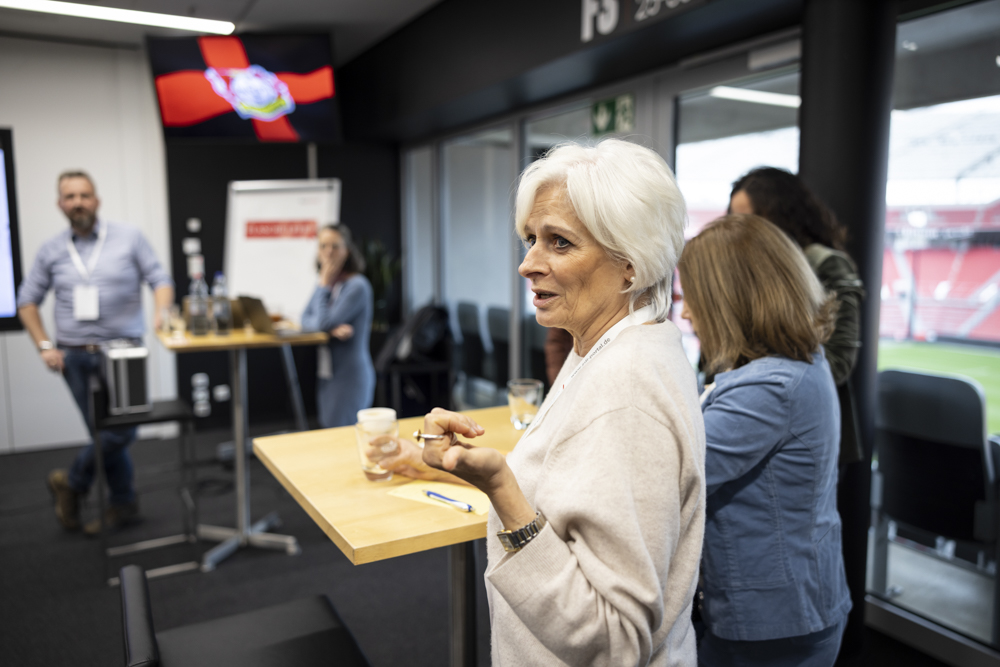 Eine ältere Frau steht und spricht an einem Tisch in einem modernen Konferenzraum mit anderen Personen. Durch die Fenster ist ein Stadion zu sehen.