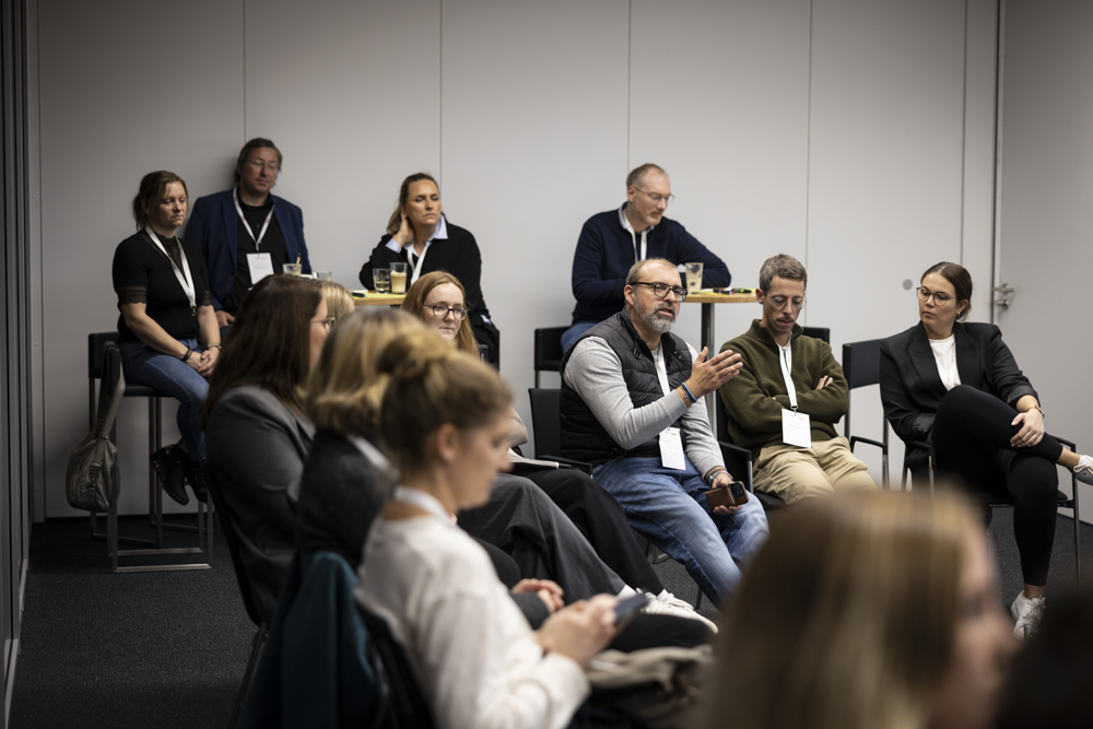 Eine Gruppe von Personen sitzt in einem Konferenzraum. Ein Mann spricht und gestikuliert mit seiner Hand, während die anderen aufmerksam zuhören.