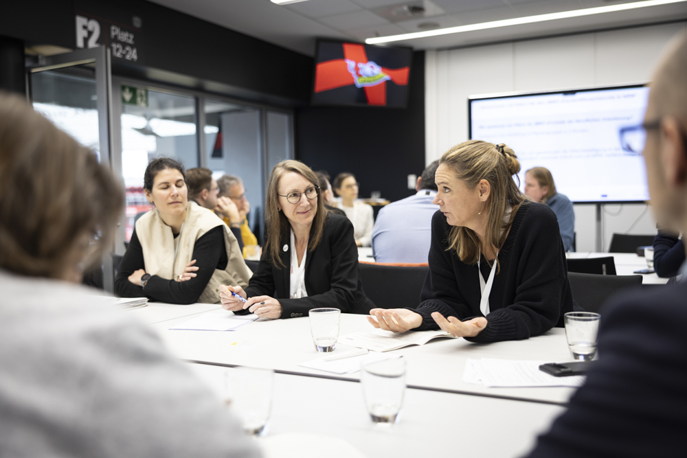 Vier Personen sitzen an einem Tisch in einem Besprechungsraum und diskutieren mit Papieren und Wassergläsern vor sich. Andere stehen dahinter.