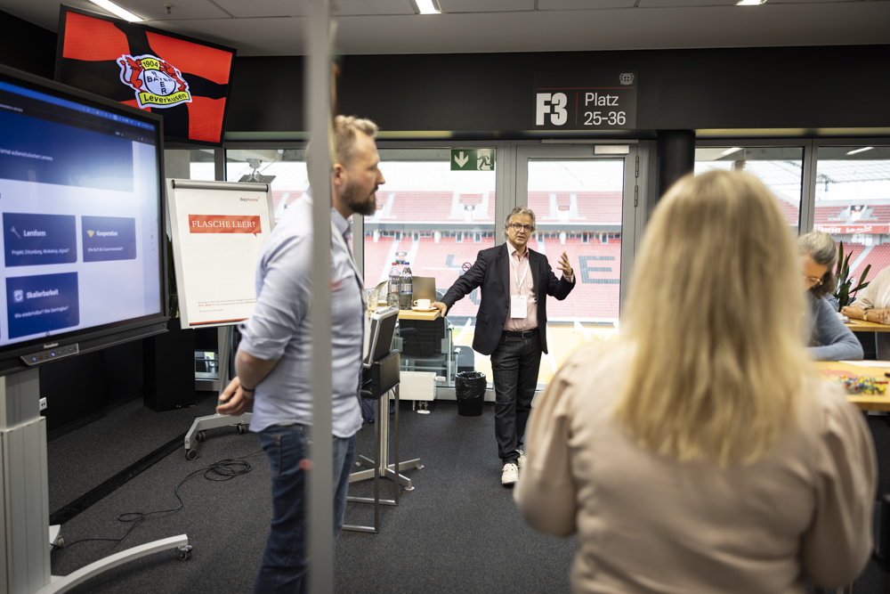 Die Leute sitzen in einem modernen Konferenzraum und diskutieren, wobei ein großer Bildschirm, ein Flipchart und Stadionsitze durch das Fenster sichtbar sind.