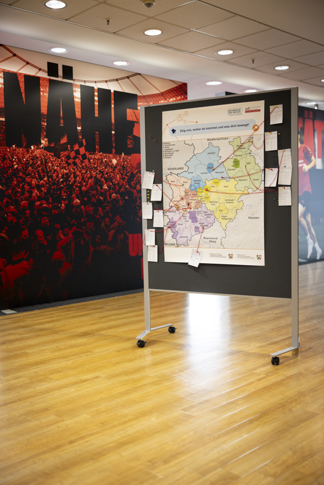 Eine mobile Anzeigetafel mit einer Karte und Papieren steht in einem Flur mit Holzfußboden und einem großen roten Wandgemälde im Hintergrund.