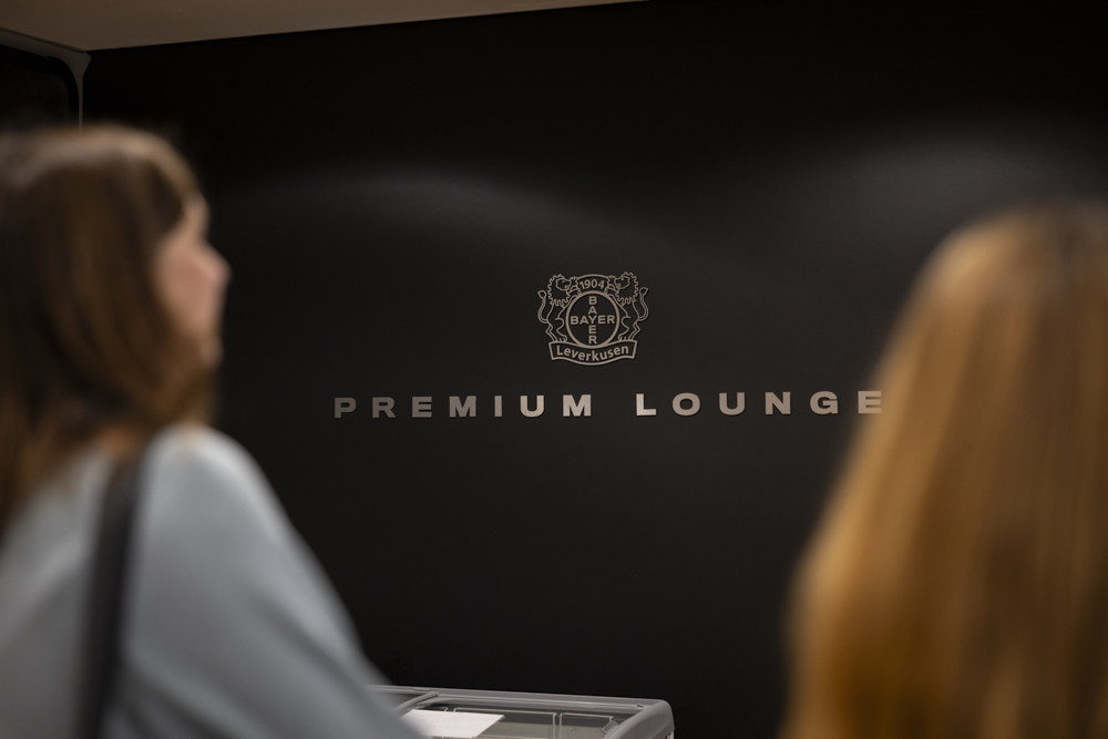 Zwei Personen stehen vor einer schwarzen Wand mit dem Logo von Bayer 04 Leverkusen und dem Schriftzug Premium Lounge" in silbernen Buchstaben.