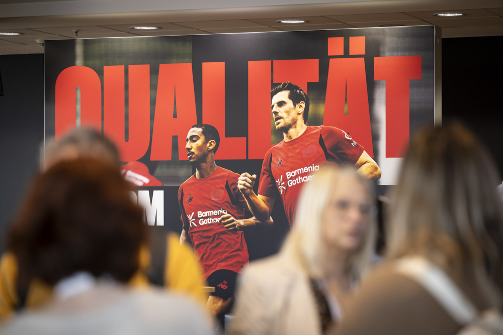 Mehrere Personen unterhalten sich vor einem Plakat, das zwei Fußballspieler in roten Trikots und das große Wort "QUALITÄT" zeigt.