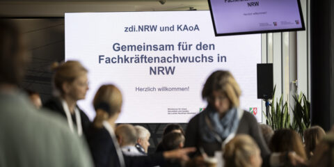 Eine Gruppe von Teilnehmern an einer Veranstaltung steht vor einem großen Bildschirm mit der Aufschrift "Gemeinsam für den Fachkräftenachwuchs in NRW".