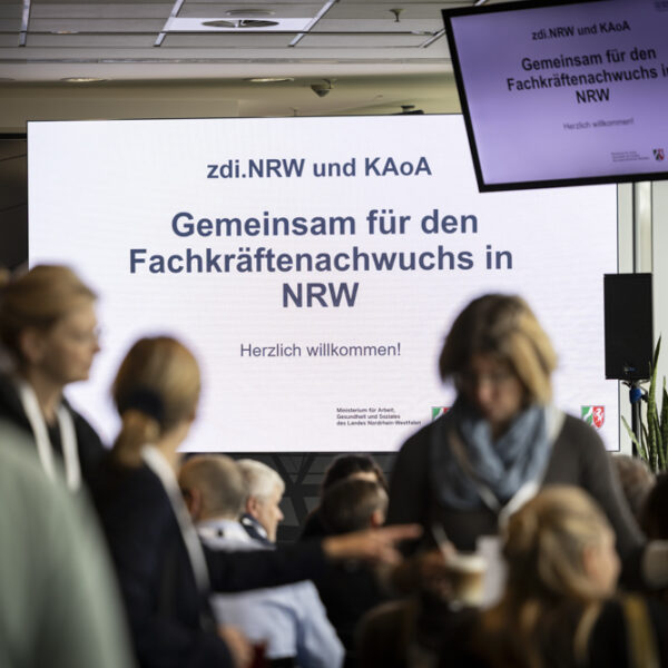 Eine Gruppe von Teilnehmern an einer Veranstaltung steht vor einem großen Bildschirm mit der Aufschrift "Gemeinsam für den Fachkräftenachwuchs in NRW".