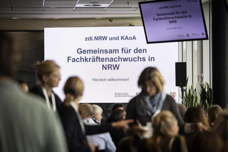 Eine Gruppe von Teilnehmern an einer Veranstaltung steht vor einem großen Bildschirm mit der Aufschrift "Gemeinsam für den Fachkräftenachwuchs in NRW".