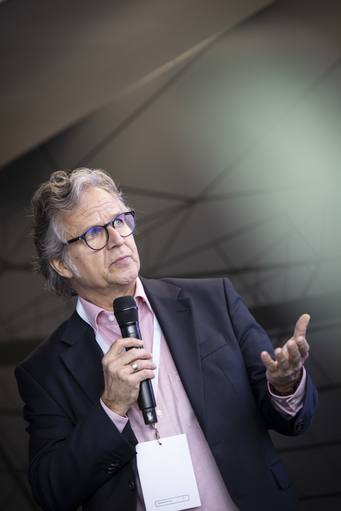 Ein Mann mit grauem Haar und Brille, gekleidet in einen Anzug und ein rosa Hemd, spricht in ein Mikrofon und gestikuliert mit einer Hand.